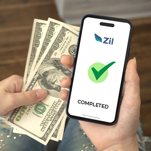 A Person Holding a Smartphone Displays the ACH Money Transfer Completed Screen with a Green Checkmark and the Zil Logo, Along with Several Dollars in the Other Hand