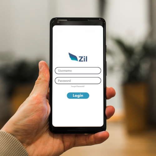 A Person Holds a Smartphone Displaying a Login Screen for an App Called "Zil." The Screen Shows Fields for "Username" and "Password," Along with a "Login" Button. The Image Portrays Zil as the Best Checking Accounts Online.