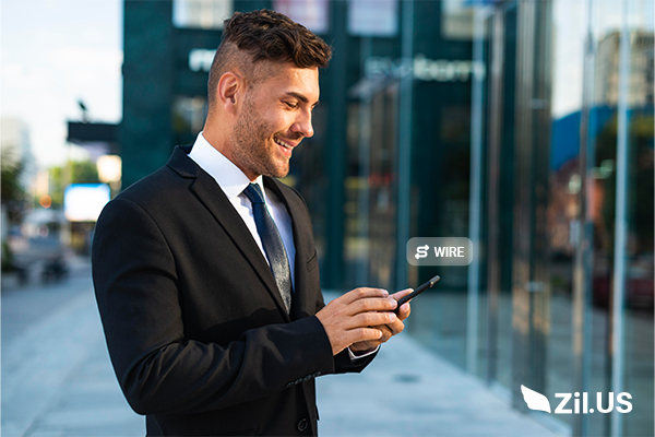 Send wire transfer quickly—young man in a formal suit holding a phone, standing on a city sidewalk near glass windows.